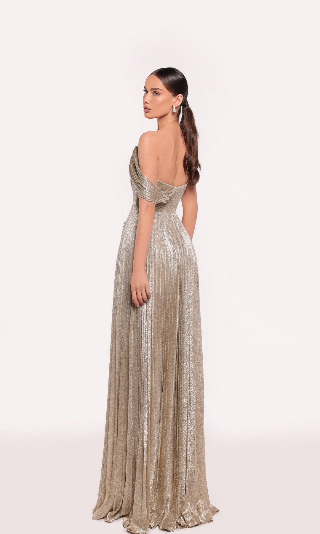A woman with her hair in a low ponytail stands facing away, wearing the gold Tarik Ediz 98725 long formal dress featuring metallic pleats and an asymmetrical neckline.